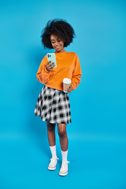 Una mujer negra se para frente a un fondo azul vibrante, sosteniendo un teléfono celular. - Foto, imagen