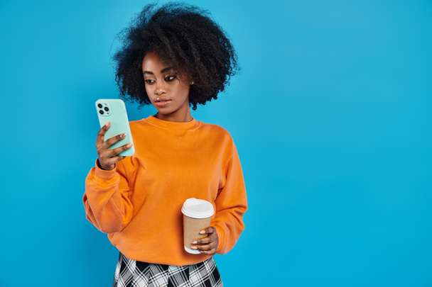 black woman stands holding a cup of coffee and a cell phone, showcasing the art of multitasking in a modern world. - Fotó, kép