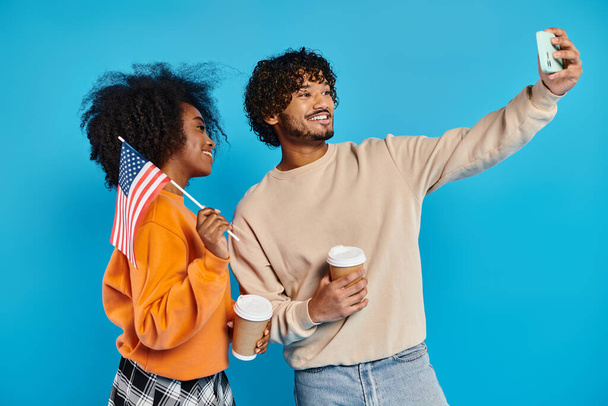 Různé rasy pár, stylově oblečený, zachycující okamžik spolu s mobilním telefonem selfie na modrém pozadí, americká vlajka - Fotografie, Obrázek