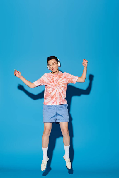 Een modieuze jongeman in trendy kleding springt vrolijk in de lucht met handen omhoog tegen een blauwe achtergrond. - Foto, afbeelding