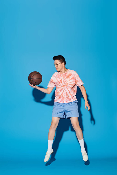 Egy divatos öltözetű, elegáns fiatalember magabiztosan tart egy kosárlabdát a jobb kezében, élénk kék háttérrel.. - Fotó, kép