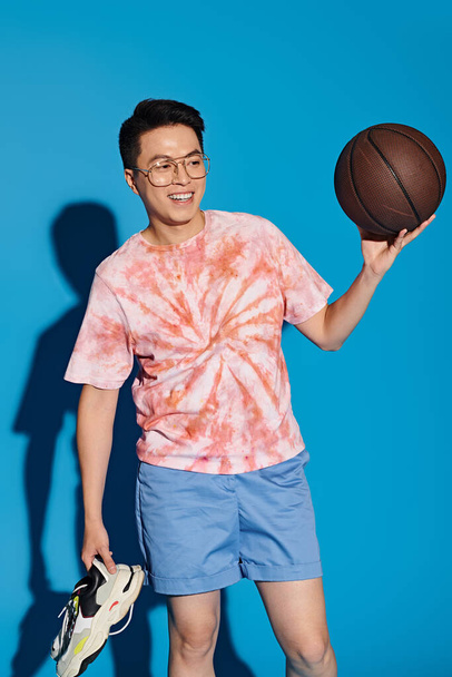 Ένας κομψός νεαρός με μοντέρνα ενδυμασία κρατάει με ενέργεια ένα μπάσκετ στο χέρι του σε ένα μπλε φόντο.. - Φωτογραφία, εικόνα