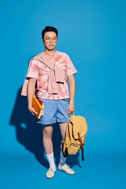Elegante joven con camisa rosa y pantalones cortos azules sosteniendo enérgicamente una bolsa amarilla sobre un fondo azul. - Foto, imagen