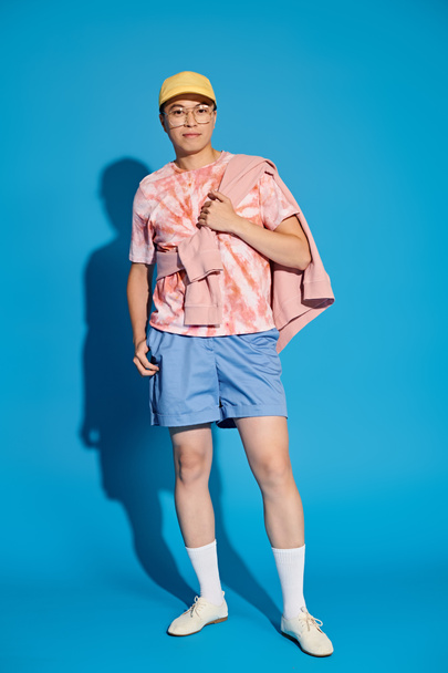 Молодой человек, стильно одетый в розовую рубашку и синие шорты, энергично позирует на синем фоне. - Фото, изображение