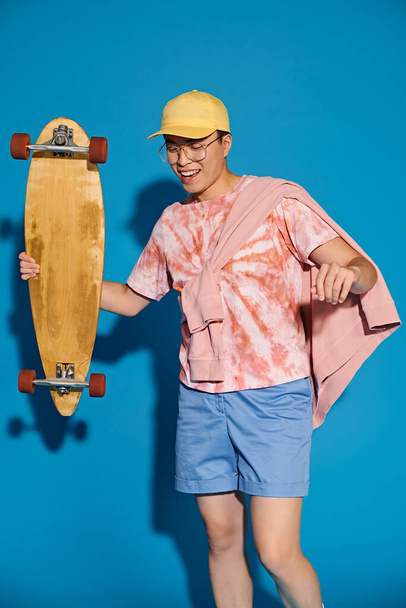 Jeune homme en tenue tendance tient avec confiance skateboard devant un mur bleu vibrant. - Photo, image