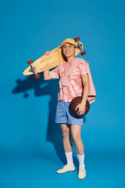 Stylový mladý muž v módní oblečení bez námahy vyváží skateboard a míč na pulzujícím modrém pozadí. - Fotografie, Obrázek