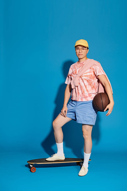 A stylish young man balances on a skateboard while holding a basketball, showcasing his skill and confidence against a blue backdrop. - Valokuva, kuva