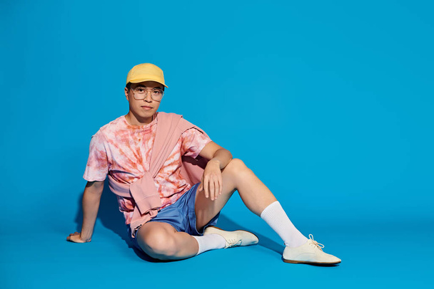 Een trendy jongeman, stijlvol gekleed, zit op de grond met een levendige gele hoed tegen een blauwe achtergrond. - Foto, afbeelding
