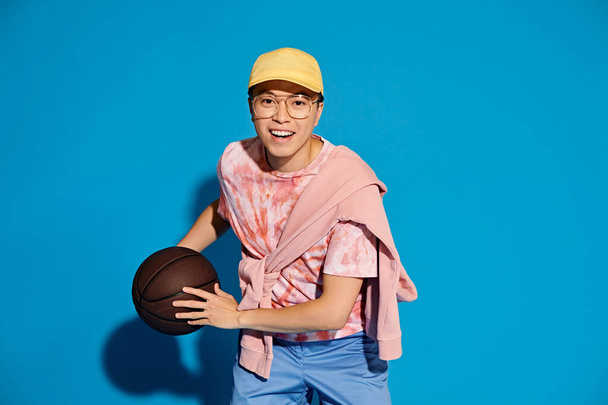 Un giovane alla moda tiene energicamente un pallone da basket nella mano destra su uno sfondo blu. - Foto, immagini