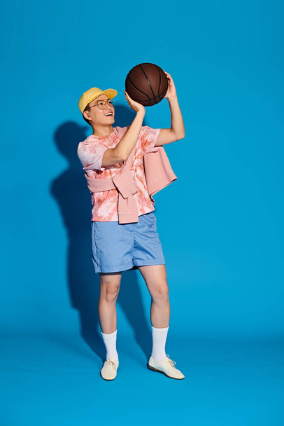 Stylish young man confidently holds a basketball in his right hand, exuding athleticism and coolness against a blue backdrop. - Fotoğraf, Görsel