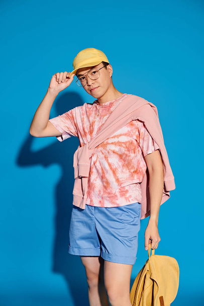 A stylish young man poses on a blue backdrop, wearing a pink shirt and blue shorts while holding a yellow bag. - Fotoğraf, Görsel