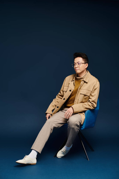 A fashionable young man in elegant attire sits on a blue chair with his legs crossed, showcasing a relaxed and stylish pose. - Foto, Bild