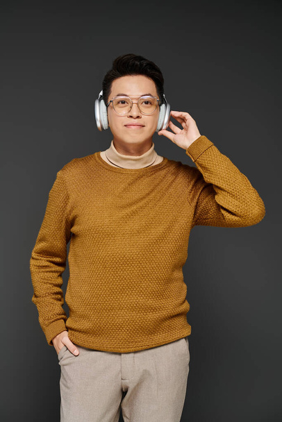 Un jeune homme à la mode dans un pull confortable écoute attentivement à travers des écouteurs élégants, respirant une confiance sereine. - Photo, image