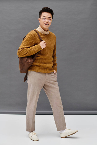 A fashionable young man strikes a dynamic pose in a brown sweater and tan pants, showcasing his elegant attire. - 写真・画像