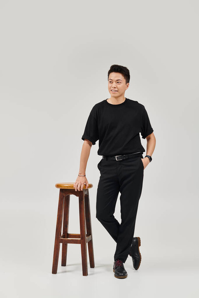 A fashionable young man in elegant attire striking a pose next to a wooden stool. - Zdjęcie, obraz