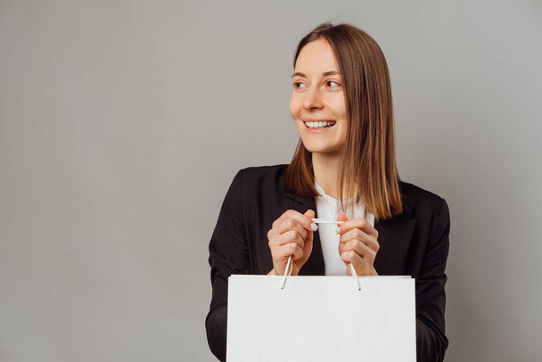Allegro giovane donna è in possesso di una shopping bag bianca, mentre guardando da parte su sfondo grigio. - Foto, immagini