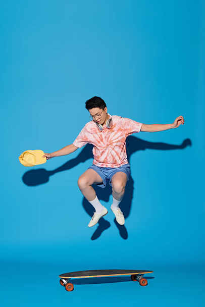 Een stijlvolle, knappe jongeman in trendy kleding springt in de lucht met een skateboard tegen een blauwe achtergrond. - Foto, afbeelding