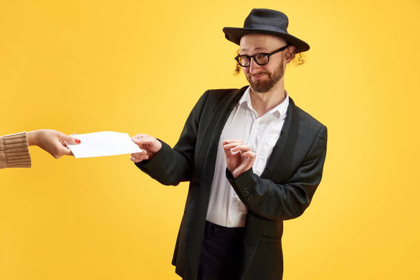 Skeptical Jewish man in hat and formal suit receiving envelope from hand against bright yellow background. Purim, business, festival, holiday, celebration Pesach or Passover, judaism, religion concept - Photo, Image