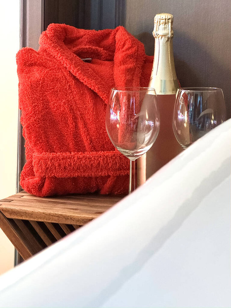A frottírvörös köntös, egy üveg pezsgő és két pohár van az asztalon a fehér fürdő mellett. A téma Valentin nap.. - Fotó, kép