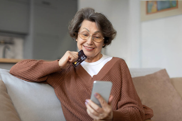 Glückliche Seniorin mit Brille genießt die Bequemlichkeit des Online-Shoppings zu Hause, schaut auf den Handybildschirm und hält Kreditkarte in der Hand - Foto, Bild