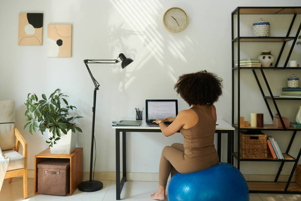 Woman sitting on fitness ball when working on laptop at home, view from behind - Photo, Image