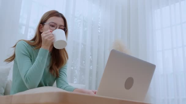 Mujer asiática joven con gafas sentado sofá de trabajo en el ordenador portátil y beber café en la sala de estar en casa, mujer de trabajo desde casa con teletrabajo, trabajo independiente, negocios y la vida digital. - Imágenes, Vídeo