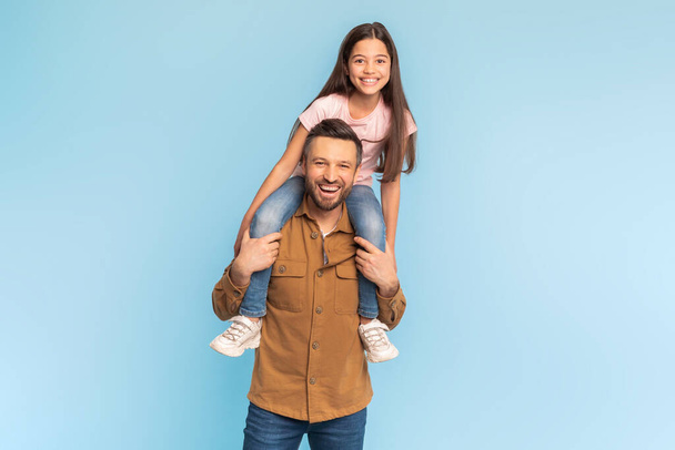 Nejlepší otec. Šťastná dceruška sedí na tatínkovi ramena usmívá se na kameru nad modrým studiem na pozadí. Pyšný tatínek pózování dávat ramena jízda na jeho půvabné dítě dívka - Fotografie, Obrázek
