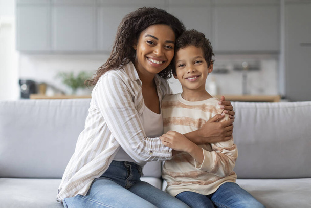 Portrait d'une jeune mère noire heureuse et de son fils préadolescent embrassant à la maison, d'une mère afro-américaine souriante et d'un petit enfant mâle se relaxant sur le canapé et s'amusant ensemble, regardant l'appareil photo, copiant l'espace - Photo, image