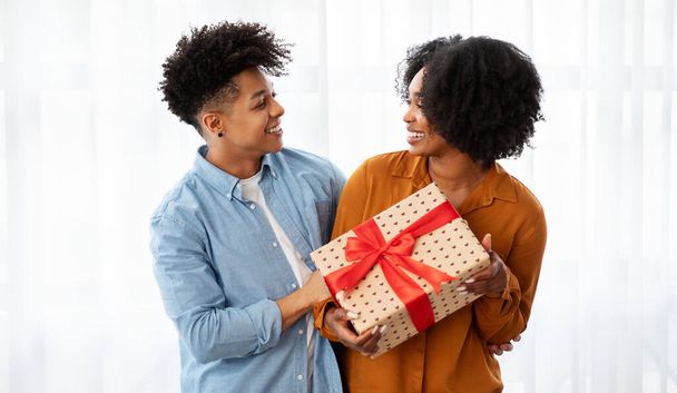 Ein fröhliches afrikanisch-amerikanisches junges Paar teilt einen besonderen Moment, als es ein wunderschön verpacktes Geschenk mit einer roten Schleife tauscht, deren Gesichter in einem hellen, luftigen Raum mit einem Lächeln erstrahlen. - Foto, Bild