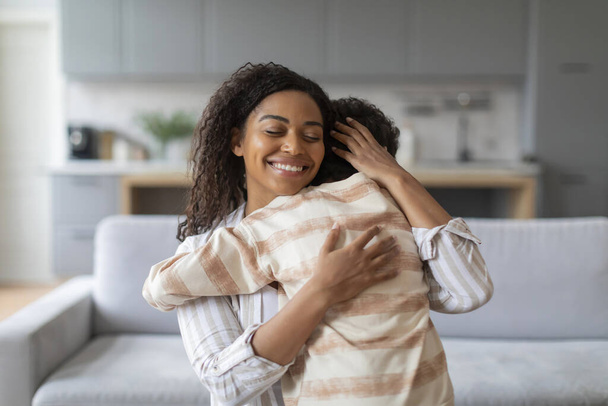 Freudige schwarze Mutter umarmt ihren preteen Sohn in liebevoller Umarmung zu Hause, afrikanisch-amerikanische Mutter und männliches Kind teilen Moment der Zuneigung, während sie zusammen in gemütlichen, gut beleuchteten Wohnzimmer entspannen, Kopierraum - Foto, Bild