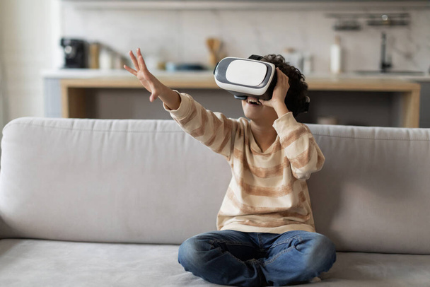 Excited black little boy wearing virtual reality headset having fun at home, african american male kid immersed in digital world sitting on sofa in bright modern living room at home, copy space - Photo, Image