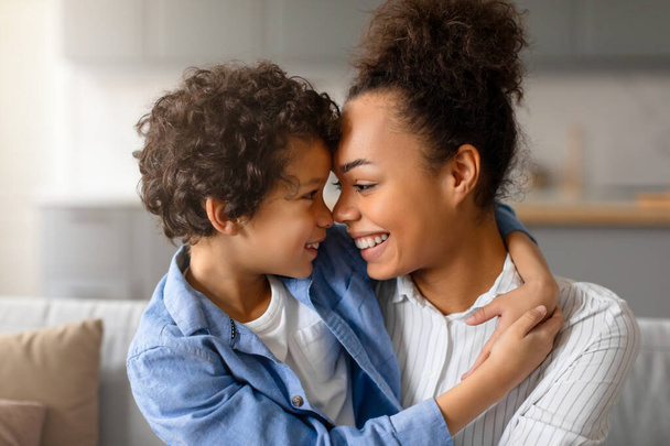 Encantada madre negra y su hijo tocando afectuosamente la frente, compartiendo un momento feliz e íntimo en su acogedora sala de estar en casa - Foto, imagen