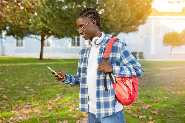Sonriente estudiante negro chico en gafas de navegación en su teléfono con auriculares listos para la música, disfruta de un día soleado al aire libre caminando con mochila en el campus universitario parque - Foto, Imagen