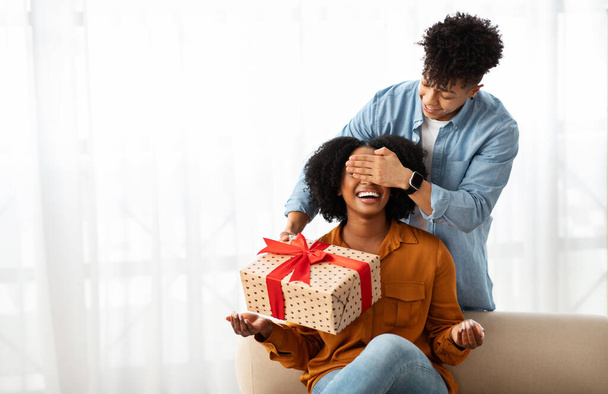 Glimlachende jonge Afro-Amerikaanse man gesloten ogen voor dame, geeft geschenkdoos, in de woonkamer interieur. Verrassing, liefde en relatie, verjaardag, jubileum, vakantieviering thuis - Foto, afbeelding