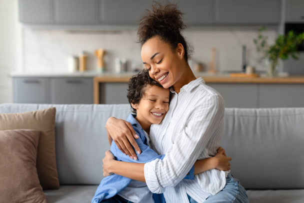 Vreugdevolle Afrikaanse Amerikaanse moeder omarmt haar zoontje met liefdevolle knuffel, het delen van moment van genegenheid in hun heldere, moderne keuken interieur - Foto, afbeelding