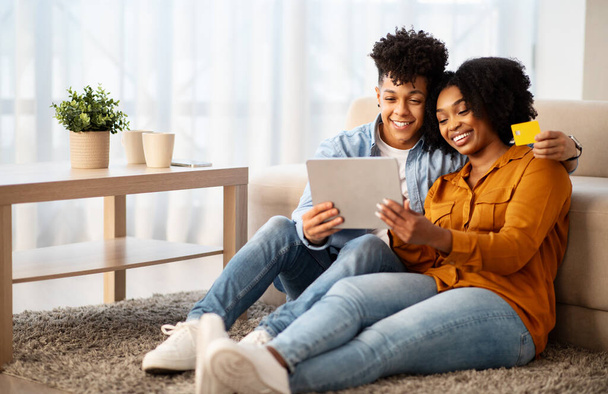 Happy young african american guy and woman shopaholics, hugs, use tablet, credit card, enjoy pay for order in living room interior. App, sale, shopping at free time at home - Photo, Image