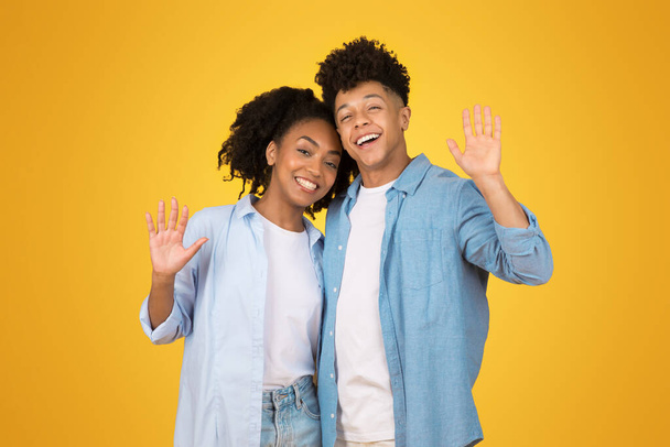 Affable jeune femme afro-américaine heureuse et homme agitant bonjour avec les mains ouvertes, à la fois des sourires brillants sportifs et tenue bleue décontractée, dans un cadre jaune joyeux - Photo, image