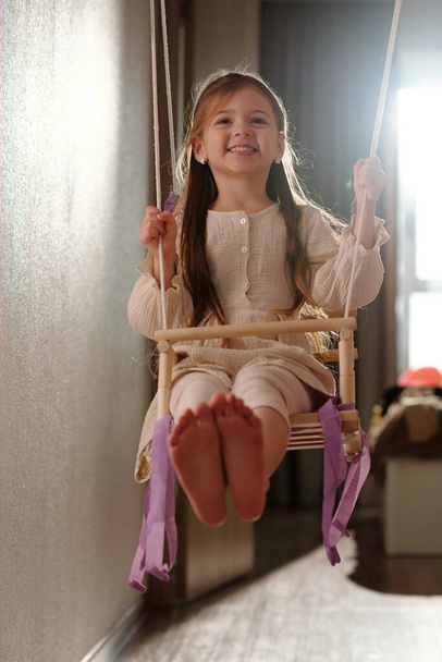 Alegre niña en vestido blanco inteligente sentado en columpio de madera delante de la cámara y sosteniendo por cuerdas mientras se divierten en casa - Foto, imagen