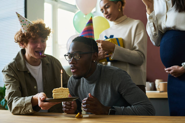 Fiatal üzletember égő gyertyát fúj egy szelet finom szülinapi tortán, miután kívánságot tett, miközben boldog kollégák között ült. - Fotó, kép