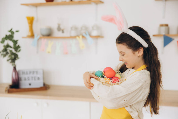 Портрет милой счастливой маленькой девочки в плюшевых кроличьих ушах и фартуке, держащей плетеную корзину, полную пасхальных творчески украшенных яиц культового цвета, стоя на кухне с праздничными украшениями - Фото, изображение
