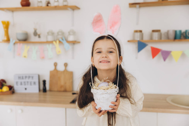 Портрет милой счастливой маленькой девочки в плюшевых кроличьих ушах и фартуке, держащей плетеную корзину, полную пасхальных творчески украшенных яиц культового цвета, стоя на кухне с праздничными украшениями - Фото, изображение