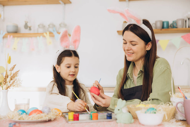Anne ve kızı yumurta boyuyor. Mutlu aile Paskalya için hazırlanıyor. Tavşan kulağı takan şirin bir kız çocuğu.. - Fotoğraf, Görsel
