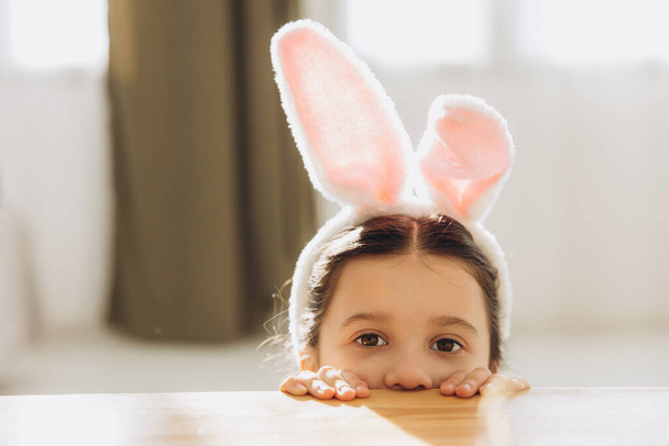 Καλό Πάσχα! Χαριτωμένο κοριτσάκι φορώντας αυτιά λαγουδάκι, ενώ κρύβονται πίσω από το τραπέζι και να έχουν κάποια διασκέδαση στην κουζίνα στο σπίτι - Φωτογραφία, εικόνα