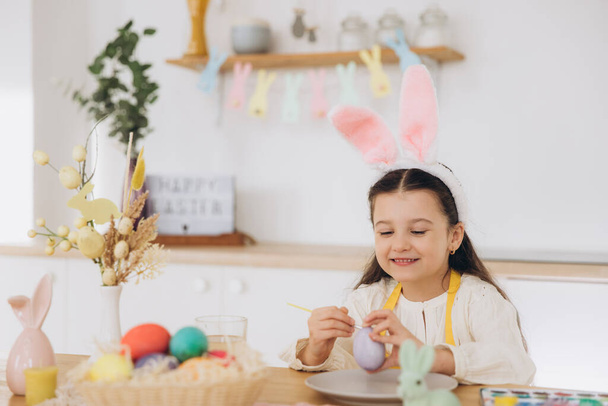 Buona Pasqua! Una bella bambina dipingere uova colorate e divertirsi. Ragazza carina che indossa orecchie di coniglio il giorno di Pasqua. - Foto, immagini