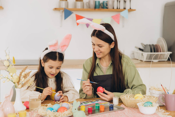 Moeder en haar dochter schilderen eieren. Gelukkige familie bereidt zich voor op Pasen. Schattig klein kind meisje draagt konijnenoren. - Foto, afbeelding