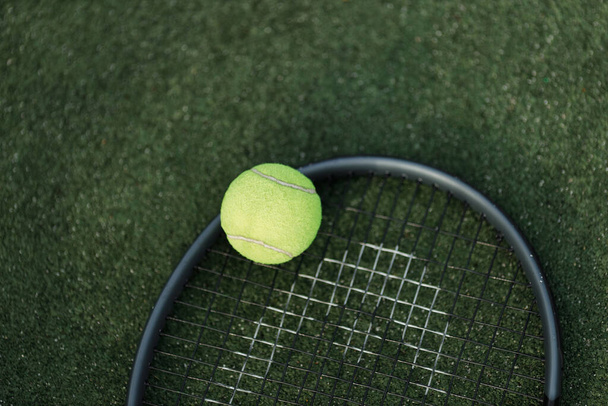 A vibrant green tennis ball rests on a black tennis racket, both set against the contrasting backdrop of a darker green court. - Photo, Image