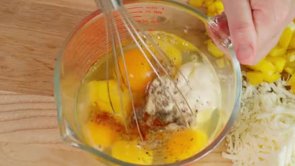 Las manos del chef en un tazón de medir vaso con un batidor mezclando huevos con especias, preparando un desayuno saludable frittata, mesa de madera, sobre un fondo de verduras. Primer plano, vista superior. - Metraje, vídeo
