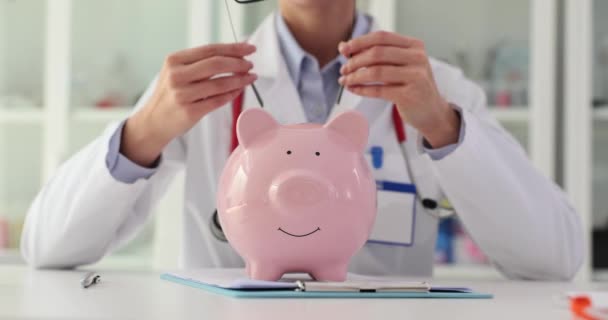 La doctora se pone anteojos en la alcancía y recomienda ahorrar dinero para los servicios de seguro médico. Tratamiento y coste de las enfermedades oculares - Metraje, vídeo