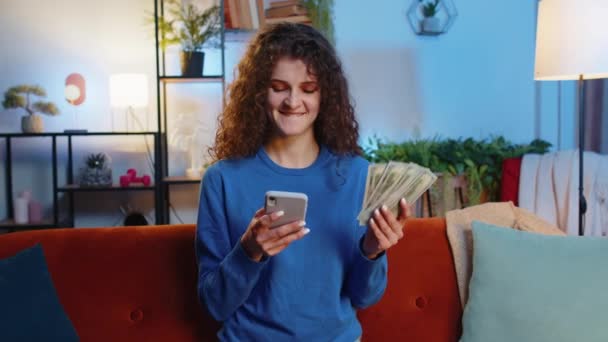Successful rich woman with curly hair holding smartphone and waving dollar bill money fan sitting on sofa at home. Happy girl winning online lottery game planning vacation calculating budget concept - Footage, Video