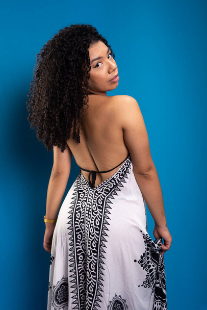 Beautiful young woman, with curly hair, standing with her back to the camera, wearing a long dress. Isolated on blue background. - Photo, Image